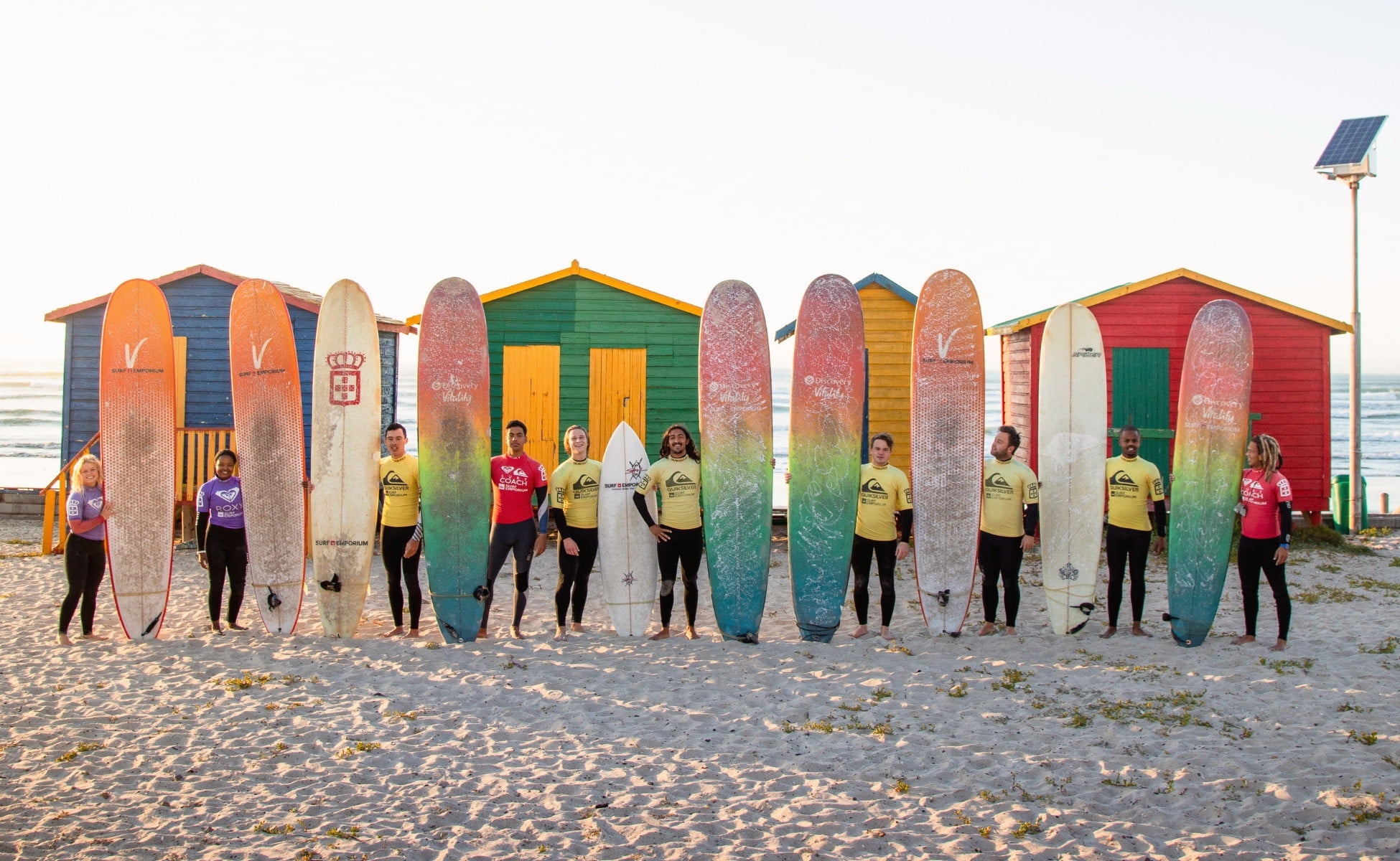 A group of young surfers stand in front ot the colourful changing cubicles on Muizenberg Beach, in False Bay, South Africa.