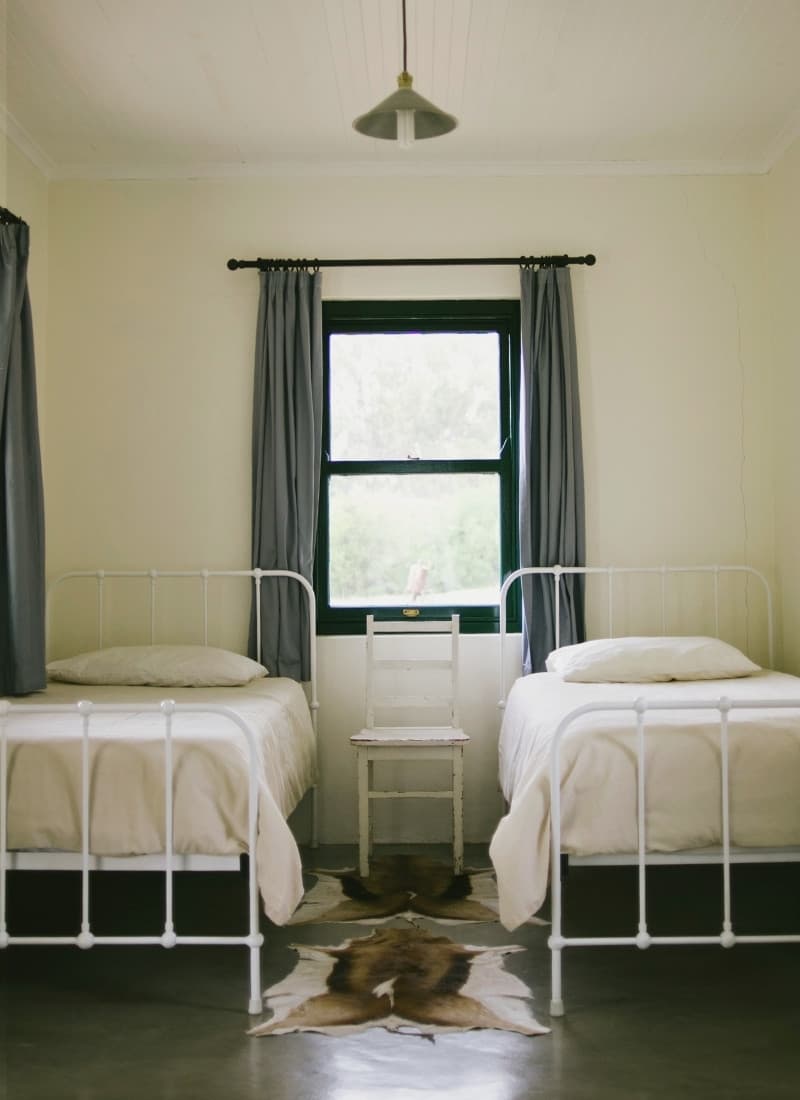 Room with two cast-iron single beds, a springbok hide between them, blue-green curtains and white walls, showing the owners’ simple, elegant style