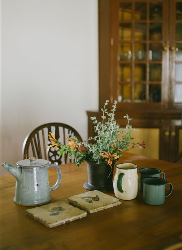 Green tin camping mugs, a cream jug, and a small ceramic vase of flowers arranged on a wooden table
