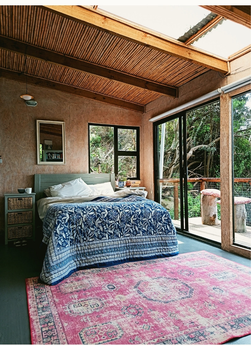 Interior of eco-cabin at Cape Point Mountain Getaway near Cape Town — bed with blue cover, red carpet and reed ceiling, with glass front opening onto balcony and afrotemperate forest