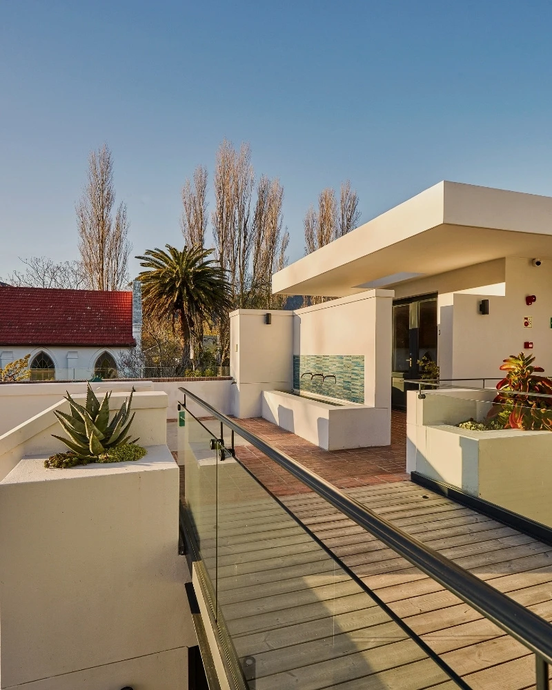 Upper balcony and water feature on the modern wing of Chapter House Franschhoek, with views of a church beyond the rooftop.