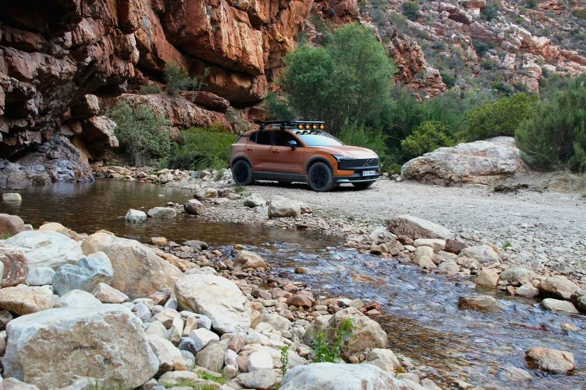 Orange Volvo EX30 parked beside the [Gamka] River in Gamkaskloof, with red sandstone cliffs behind