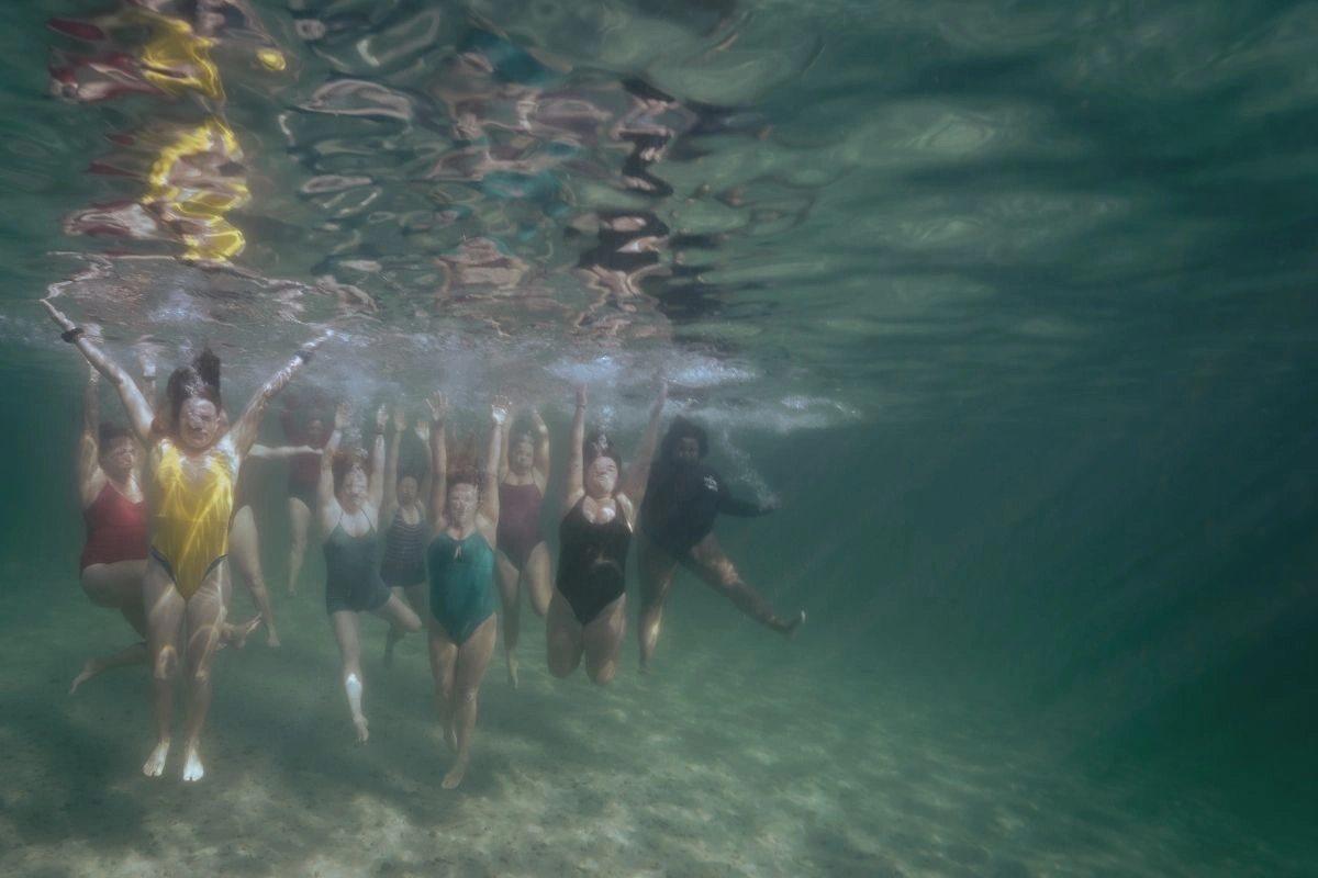 A group of women in swimming costumes underwater with their arms raised during a Helen Walne group shoot