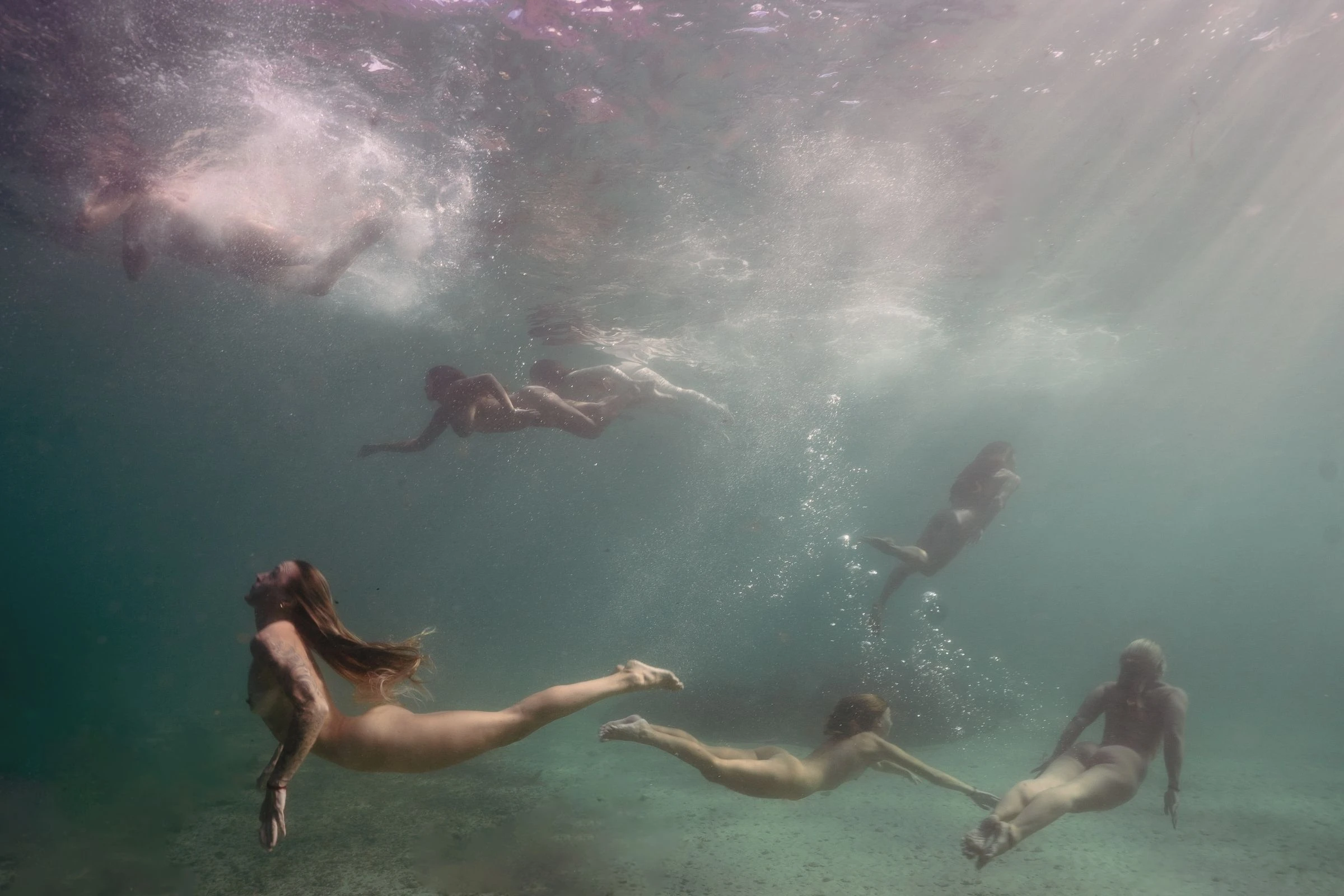 Women swimming naked up towards the surface from the sandy seabed, trailing bubbles, in a Cape Town kelp forest
